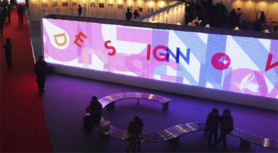 A huge screen is seen in a dark exhibition space, in front of it, people are sitting on a bench